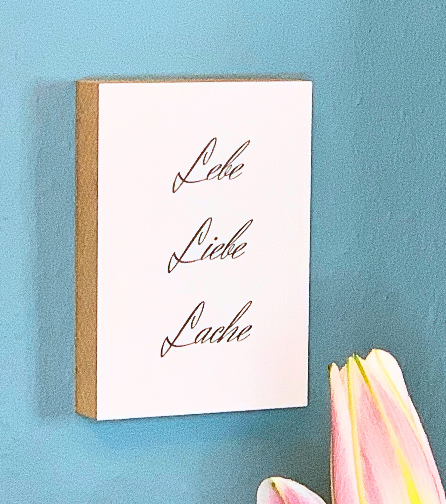 Holzbild mit schwarzer Schrift auf weißem Hintergrund "Lebe. Liebe Lache" auf einer blau-grauen Wand mit einer Blüte im Anschnitt.