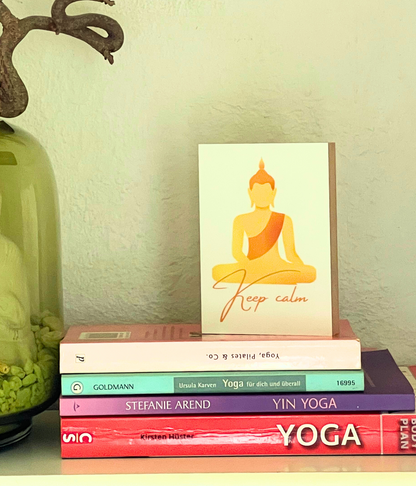 Holzbild mit dem Motiv "Keep calm" steht auf mehreren liegenden Yoga Büchern, im Anschnitt eine grüne Vase mit Zweigen