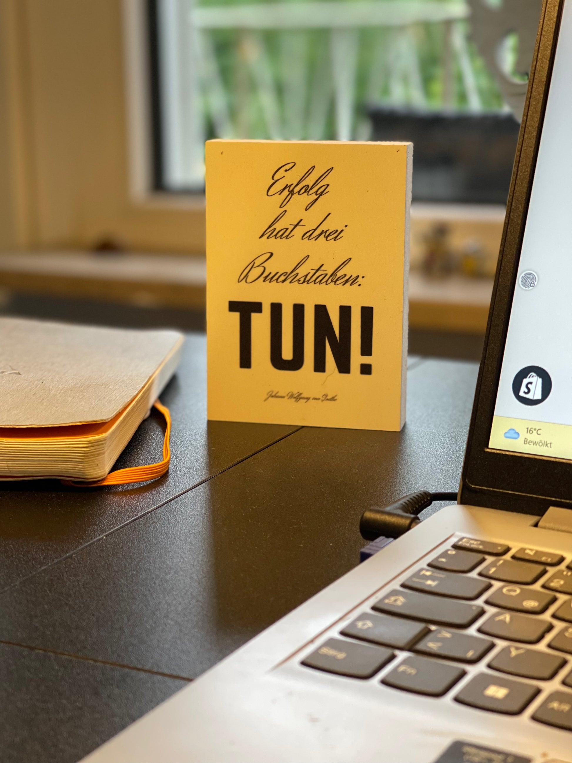 Holzbild mit dem Text "Erfolg hat drei Buchstaben:Tun!" steht auf einem S>chreibtisch neben Laptop und Kladde.